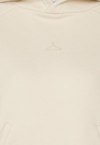 Sudadera con capucha beige hecha de tela suave con un bolsillo frontal, que presenta un logo texturizado de perchero en el centro. Diseño sencillo y detalles mínimos.