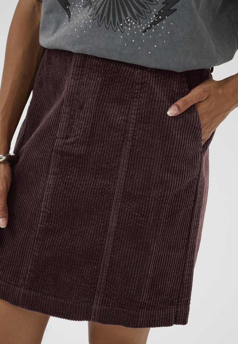 Jupe en velours côtelé violet présentant des côtes verticales, des poches latérales et une coupe droite. Le tissu a une texture douce et une finition lisse.