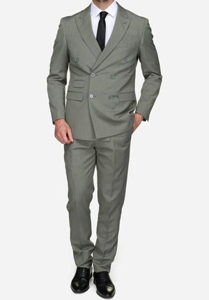 Homme portant un costume croisé gris clair avec un pantalon assorti, une chemise blanche, une cravate noire et des chaussures habillées noires, debout avec une main dans la poche.