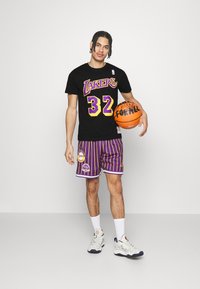 Camiseta negra con "LAKERS" y el número 32 en púrpura, combinada con pantalones cortos a rayas púrpuras con logotipos. Sosteniendo un balón de baloncesto naranja.