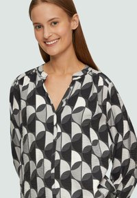 Blouse à motif noir et blanc avec un décolleté en V, présentant des formes arrondies et un tissu texturé. Manches longues avec épaules froncées.