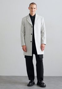 Light gray wool overcoat with a notched collar, three buttons, and two front pockets, paired with black pants and shiny black shoes.