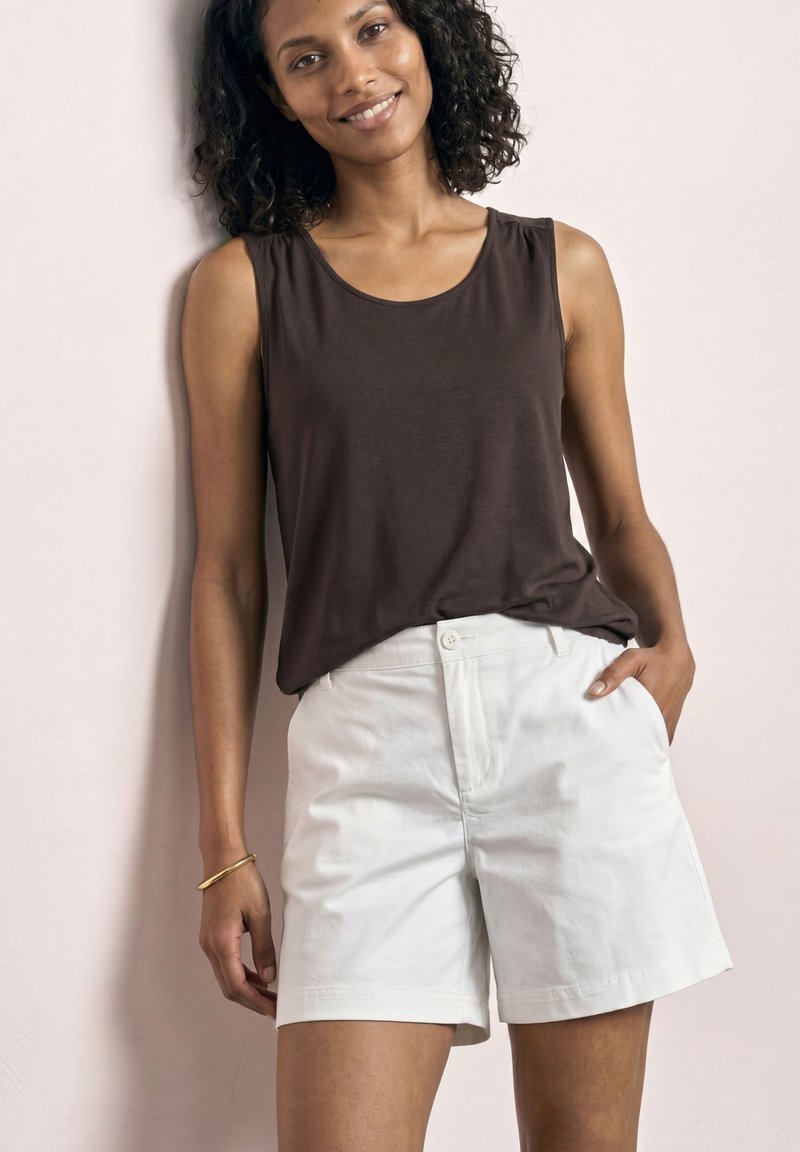 Femme souriante, portant un haut sans manches marron foncé et un short blanc taille haute, debout avec une main dans la poche contre un mur pâle.