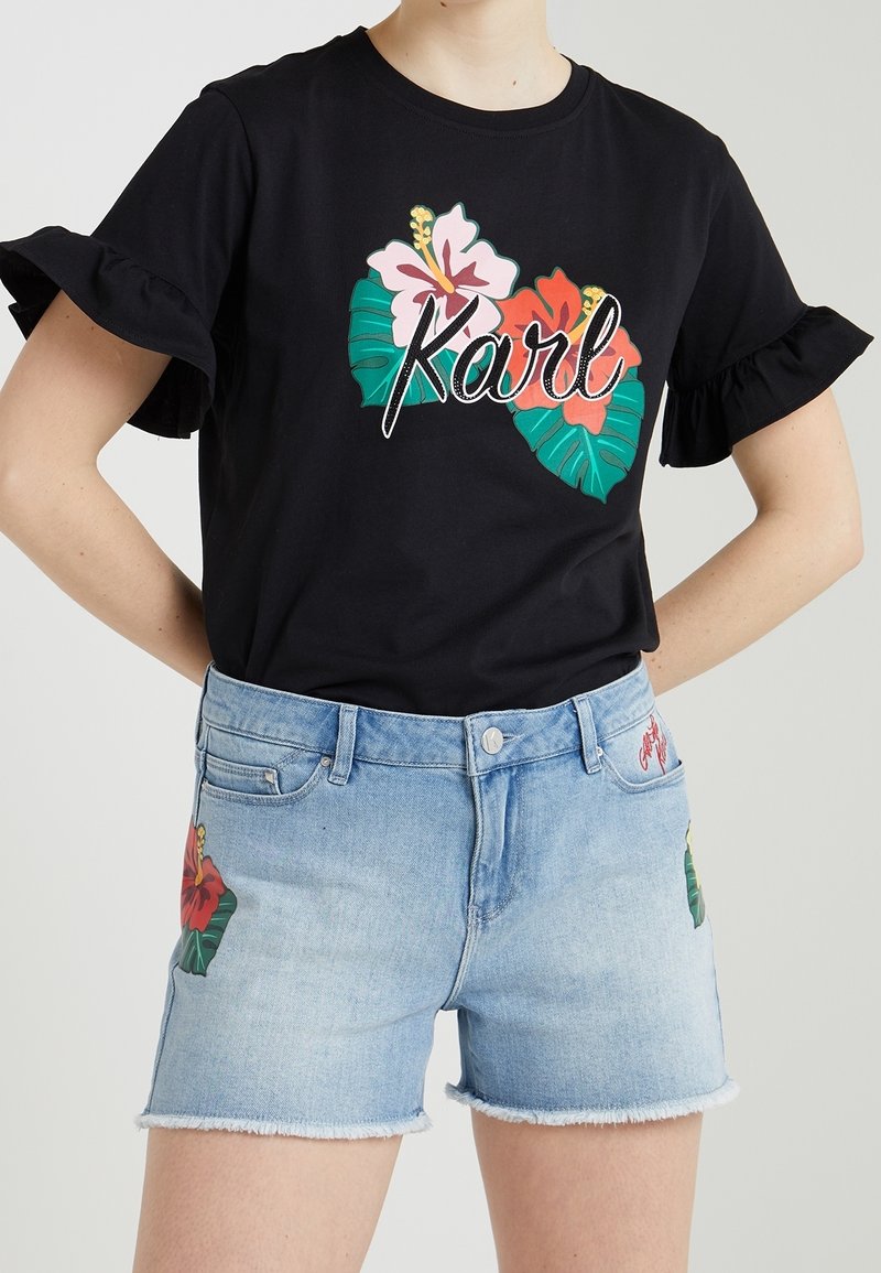 Camiseta negra con mangas rizadas que presenta gráficos coloridos de flores y hojas, combinada con pantalones cortos de mezclilla azul claro con diseños florales similares.