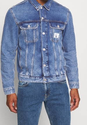 Uomo che indossa una giacca di jeans azzurro chiaro con bottoni e tasche con patta e jeans blu scuro su uno sfondo neutro.