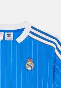 Maillot de football bleu à rayures avec col blanc. Présente le logo brodé du Real Madrid et trois rayures blanches et orange sur l'épaule.