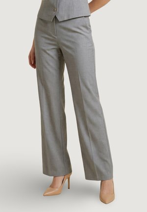 Mujer que lleva pantalones a medida de color gris claro y zapatos de tacón beige, de pie con una mano relajada a su lado.