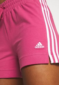 Vaaleanpunaiset urheilushortsit, jotka on valmistettu puuvillasta ja joissa on valkoiset korostusraidat sekä Adidas-logo. Sisältää joustavan vyötärön ja sivuaukot.