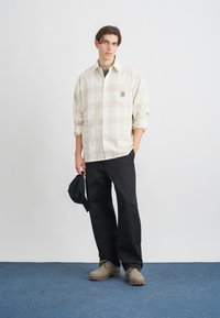 Φωτεινό καρό πουκάμισο με κουμπιά και τσέπη στο στήθος, μαύρο παντελόνι και μπεζ παπούτσια. Το μοντέλο κρατά μια μαύρη κάππα. Μαλακές υφές και άνετη γραμμή.