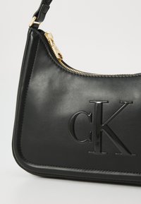 Crna kožna torba za rame sa zlatnim zatvaračem, sa utisnutim "ck" logom. Glatka tekstura, zakrivljeni oblik i detalji kontrastnog šivanja.