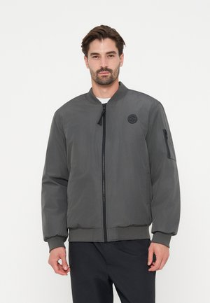 Mann mit dunklen Haaren, der eine graue Bomberjacke mit Reißverschluss, gerippten Bündchen und einem kleinen runden Logo auf der Brust trägt.