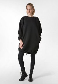 Musta ylisuurinen mekko, jossa on teksturoitu kangas, pyöreä pääntie ja elastinen kiristin helmassa, yhdistetty mustiin legginsseihin ja nauhakenkäisiin sotilaskenkään.