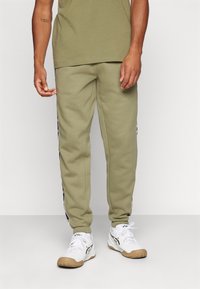 Joggers verde oliva hechos de un tejido suave con franjas laterales y puños. Combinados con zapatillas deportivas blancas con suela de goma.