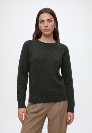 Femme aux longs cheveux bruns portant des boucles d’oreilles dorées, un pull en maille vert foncé et un pantalon beige, debout devant un fond clair uni.