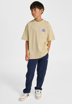 Beige bomulds-T-shirt med en blå logo, kombineret med marineblå træningsbukser og hvide sneakers med sorte striber. Enkel og afslappet påklædning.