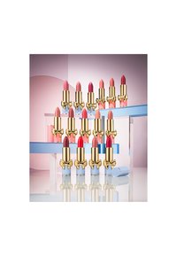 Collection of lipsticks in gold tubes with bow accents, featuring various shades of pink, red, and nude, displayed on blue geometric stands.
