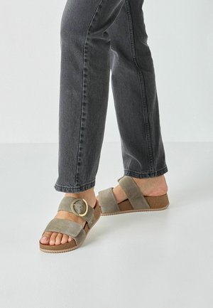 Graue Wildleder-Sandalen mit breitem Riemen und runder Metall-Schnalle, ausgestattet mit einer Korksohle und einem überkreuzten Design. Kombiniert mit dunklen Jeans.