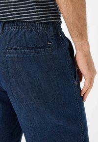 Pantaloni in denim blu scuro con vita elastica e texture liscia, tasca laterale, tasca posteriore con patch e un piccolo etichetta "Braxx" vicino alla vita.