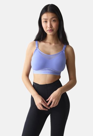 Jeune femme aux longs cheveux foncés portant un soutien-gorge de sport bleu clair et un legging noir taille haute, debout devant un fond uni.