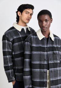 Zwei Personen tragen karierte Hemden in Grautönen mit weißen Streifen. Jedes Hemd hat einen hellen, flauschigen Kragen und vordere Knöpfe.