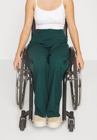 Personne assise en fauteuil roulant portant un haut blanc sans manches, un pantalon vert foncé et des chaussures beiges sur un fond blanc uni.