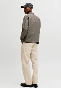 Braunes kariertes Hemd mit dunklem Kragen, cremefarbene Hose, schwarze Schuhe und schwarze Mütze. Sichtbare Nähte und Taschen an der Hose.