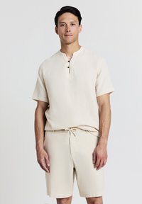 Uomo in piedi con una camicia a maniche corte beige con bottoni e pantaloni corti coordinati con cordoncino, su uno sfondo bianco semplice.