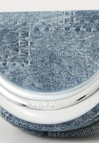 Prim-plan al unui material denim albastru cu detalii de cusătură albă și o placă metalică argintie gravată cu "REPLAY" pe călcâiul unei adidaș.