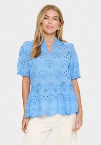 Blusa di cotone azzurro chiaro con maniche corte a palloncino, orlo scallop e dettagli in ricamo a occhio. Presenta una scollatura a V e una vita arricciata.