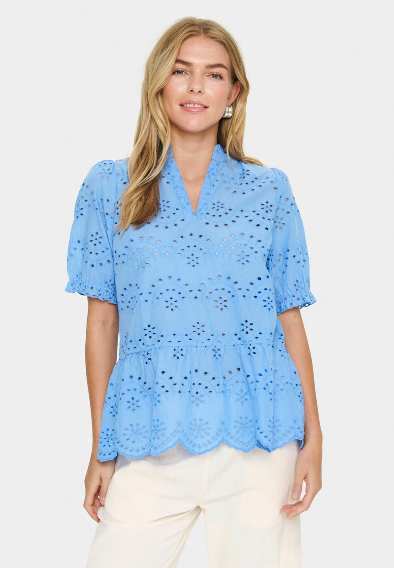 Blusa di cotone azzurro chiaro con maniche corte a palloncino, orlo scallop e dettagli in ricamo a occhio. Presenta una scollatura a V e una vita arricciata.
