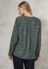 Blouse à manches longues avec une base noire ornée de motifs de feuilles turquoise et blanches. Le tissu semble léger avec une coupe décontractée et un pli au dos.