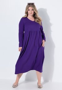 Langes lila Kleid mit V-Ausschnitt und geraffter Taille. Weicher, fließender Stoff reicht bis zur Mitte der Waden und wird mit metallischen flachen Schuhen kombiniert.