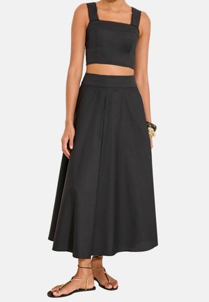 Femme portant un haut court noir sans manches avec bordure en dentelle et une jupe midi noire large taille haute avec des sandales plates.