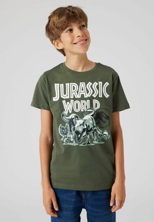 Groene katoenen t-shirt met 'Jurassic World' graphic featuring dinosaurussen. Korte mouwen, ronde hals, casual pasvorm. Blauwe spijkerbroek zichtbaar.