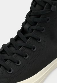 Sneaker high-top nera con materiale testurizzato, lacci tondi e occhielli neri. La suola è di colore crema ed è leggermente rialzata.