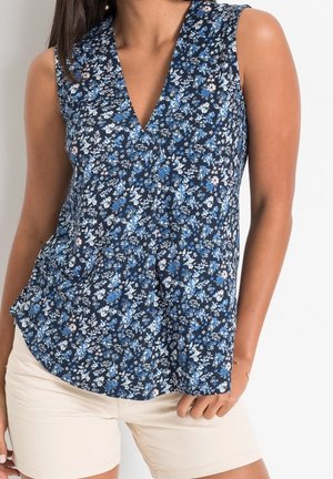 Femme portant un chemisier sans manches bleu marine à motifs floraux avec col en V et un short beige clair, debout devant un fond uni clair.