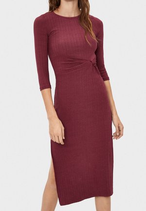 Robe en tricot côtelé bordeaux avec des manches trois-quarts, détail latéral tordu et fente haute jusqu'à la cuisse, présentant une silhouette ajustée.