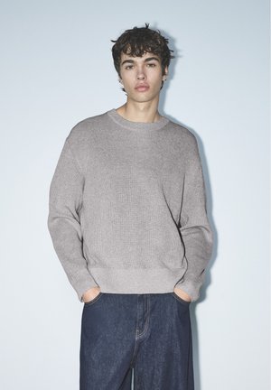 Un jeune homme se tient debout, les mains dans les poches, vêtu d’un pull en maille gris clair et d’un jean bleu foncé, sur un fond uni.
