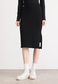 Femme portant une jupe noire côtelée longueur genou avec une fente latérale, des chaussettes blanches et des baskets blanches, debout sur un sol gris clair.