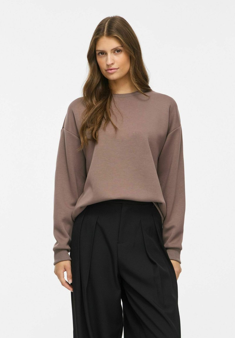 Jeune femme aux longs cheveux bruns portant un sweat-shirt marron ample et un pantalon plissé noir taille haute, debout devant un fond uni.