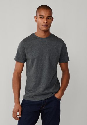 Uomo che indossa una maglietta a maniche corte di colore grigio scuro unita e jeans blu scuro, in piedi con una mano in tasca contro uno sfondo grigio chiaro.