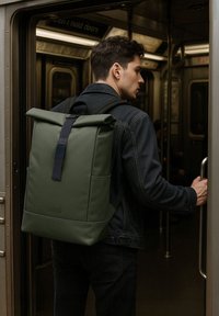 Zaino verde a rotolo realizzato in materiale liscio, con una tracolla nera. Il design include tasche laterali e una forma rettangolare elegante.