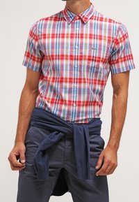 Koszula w kratę z krótkim rękawem w kolorze czerwonym, niebieskim i białym z kołnierzykiem typu button-down oraz logo Lacoste. Połączona z ciemnoniebieskimi spodniami i swetrem zawiązanym w talii.