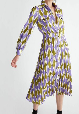 Kvinde iført en plisseret kjole med knaplukning, lange ærmer, i lilla, olivengrøn og hvidt geometrisk mønster, med to brystlommer.