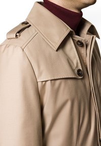 Beżowy trencz z wyprofilowanym kołnierzem, guzikami oraz gładkim, teksturowanym materiałem. Posiada pagony na ramionach i dopasowany krój.