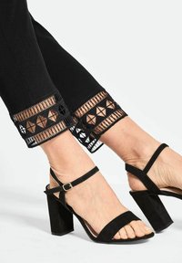 Zwarte broek tot de enkels met geborduurde, textuurvolle zomen, aangevuld met zwarte suède hoge hakken met enkelbandjes.