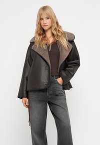 Brun faux læderjakke med en brun shearlingkrave, åben front og afslappet pasform. Kombineret med mørkegrå viddebenede jeans.