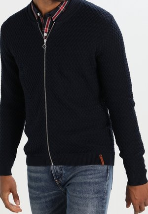 Mand iført en marineblå tekstureret zip-up sweater over en rød og sort ternet skjorte med blå jeans, vist fra hals til hofter.