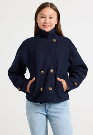Giacca doppiopetto blu navy con colletto alto e bottoni dorati decorativi; la texture simile alla lana e le tasche laterali valorizzano il design.