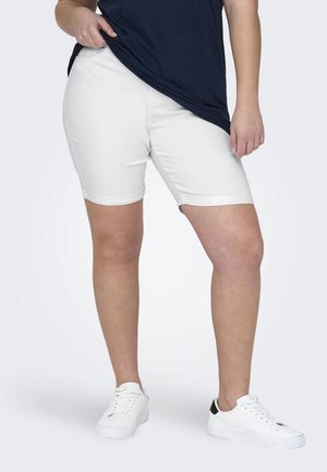 Vrouw draagt knielange witte short, donkerblauw shirt en witte sneakers, staat met één hand het zoom van het shirt vasthoudend.